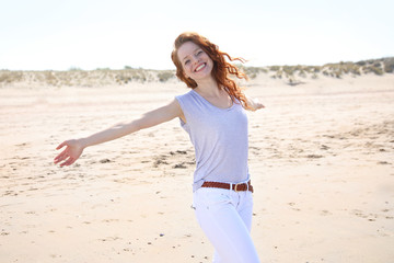 Hübsche rothaarige Frau steht mit ausgebreiteten Armen an einem Strand und lacht