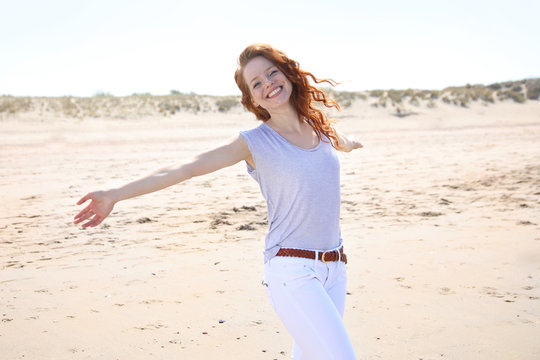 H&uuml;bsche rothaarige Frau steht mit ausgebreiteten Armen an einem Strand und lacht
