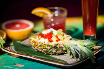 Salad with fresh vegetables, chicken and quail eggs in pineapple among refreshing cocktails from tropical fruits on a colorful table in the sun.