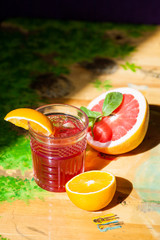 Red sweet refreshing cocktail with ice, grapefruit and orange lie on a wooden yellow table in the sun.