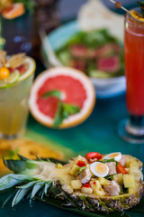 Salad with fresh vegetables, chicken and quail eggs in pineapple among refreshing cocktails from tropical fruits on a colorful table in the sun.