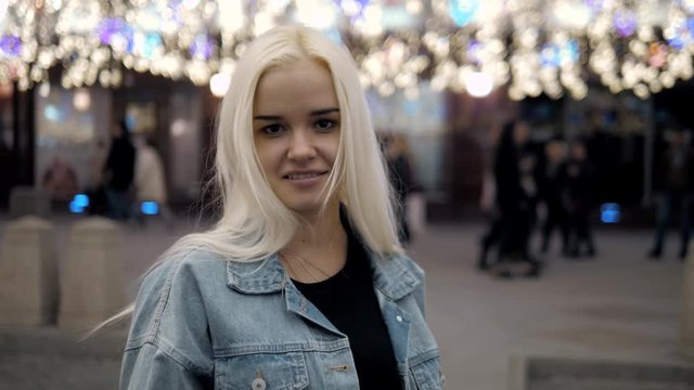 Night portrait of blonde girl using mobile phone on street, happy student model girl lights city street