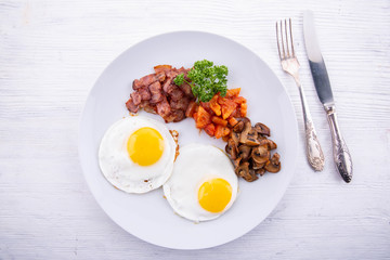 Eggs in the form of fried eggs with tomatoes, bacon, mushrooms and greens on a white wooden background