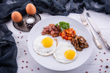 Fried eggs, boiled eggs, tomatoes, champignons, sausages, greens, salt, spices are served on a white plate for breakfast
