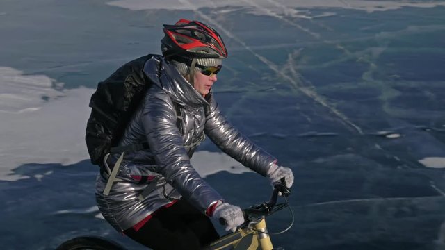 Woman is riding bicycle on the ice. The girl is dressed in a silvery down jacket, cycling backpack and helmet. Shooting with a quadrocopter drone. Ice of the frozen Lake Baikal. The tires on the