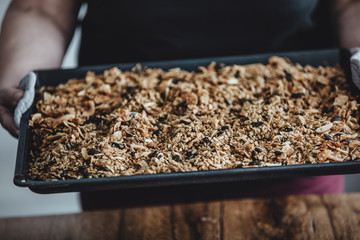 Woman holding a baking tray with freshly baked homemade granola. Healthy vegan snack easily made at home. Visible body parts of an elderly woman.
