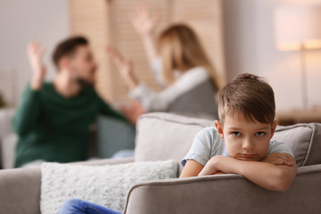 Little unhappy boy sitting in armchair while parents arguing at home