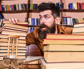 Deadline concept. Teacher or student with beard sits at table with glasses, defocused. Man on shocked face between piles of books, while studying in library, bookshelves on background.