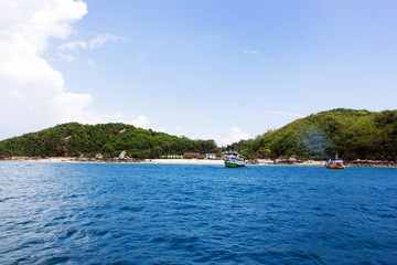 The beach on the island is surrounded by blue sea, light sky and foggy clouds. There is a boat and a resort on the island.