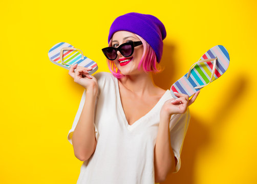 Young girl with pink hair in purple hat and sunglasses holding a flip flops shoes on yellow background