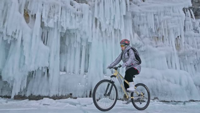 Woman is riding bicycle near the ice grotto. The rock with ice caves and icicles is very beautiful. The girl is dressed in silvery down jacket, cycling backpack and helmet. The tires on covered with