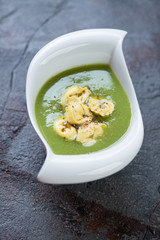 Bowl of cream-soup with broccoli topped with tortellini, vertical shot on a weathered asphalt background, selective focus
