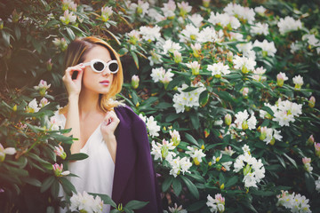 Obraz na płótnie Canvas Young redhead girl in white sunglasses near flowers at springtime blossom garden.