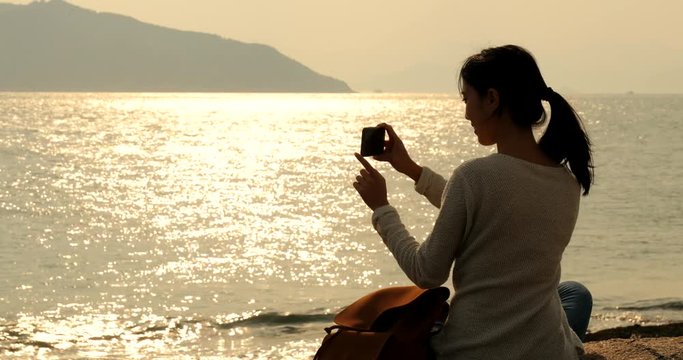 Woman enjoy the view of the sea during sunset time and taking photo on cellphone