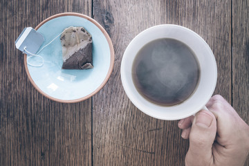 hand holding cup of steaming hot tea on old rustic wooden table with tea bog on saucer