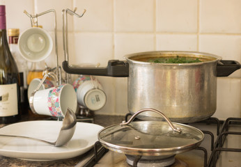 Full pot of freshly brewed soup is on a stove in a kitchen. Ready for eat. Open lid, ladle, plate, bottle of wine and other kitchen utensils - all are on a table nearby the pot.  Finished cooking. 