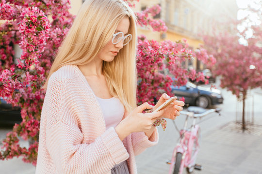 Trendy blond woman in eyeglasses traveler with bicycle using mobile phone near pink bossom apple tree at city street in spring time. Tourism, travel and new impression concept.