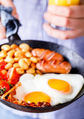 Traditional English Breakfast in the Frying Pan .Egg in the Heart shape. Valentine's Day. Festive Food in the hands of a young man Background.Sausages, Bacon, Beans,Orange juice