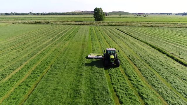 Aerial footage flying backwards in front of tractor cutting grass the cut grass will dry and then later picked up and used as animal fodder animal feed used for domesticated livestock in winter 4k