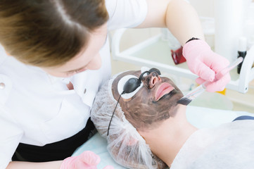 Obraz na płótnie Canvas The cosmetologist in pink gloves with a brush applies a carbon mask for peeling on the face of a young girl in a cosmetology room. The concept of cosmetology services and self-care. The concept of
