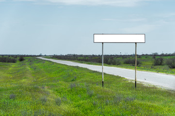 Empty old road sign for the name of a city or village populated area near the road in the countryside against a blue sky and white clouds surrounded by green grass