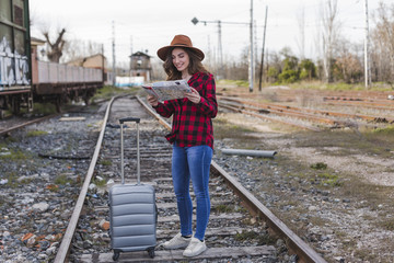 young beautiful woman wearing casual clothes, walking by the railway with suitcase and a map, she is smiling. Outdoors lifestyle. Travel concept.