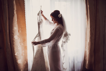 Beauty bride is holding her dress near window in the morning