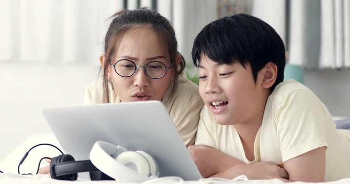 Cute asian preteen boy rest on bed with his sister and using on laptop computer with smile face at home.