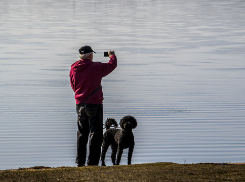 The Old Man Stay On The Beach Of Lake With Poodle Dog For Making Photo Of The View