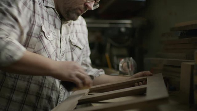 Slow motion tilt down to man sanding wood frame in workshop / Provo, Utah, United States
