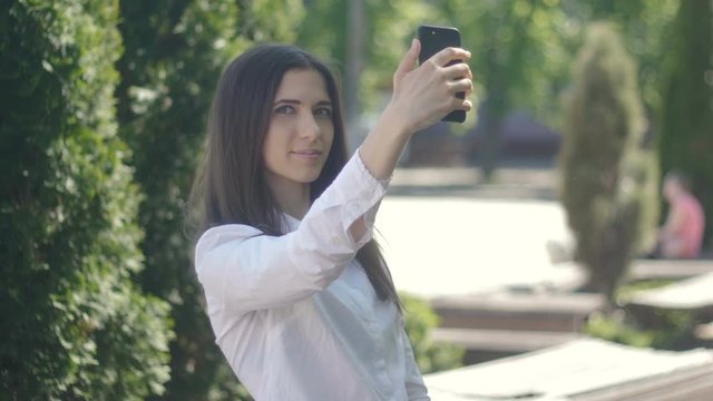young woman is photographing herself on a smartphone in the daytime outdoors. The girl makes a selfi afternoon in the city park