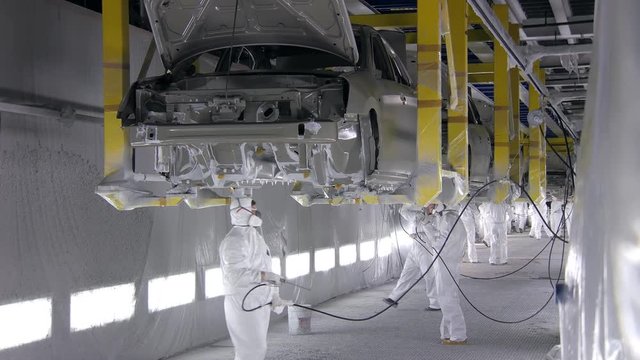 people are covering bottom of automobiles on conveyor belt , men are dressed in protective suits
