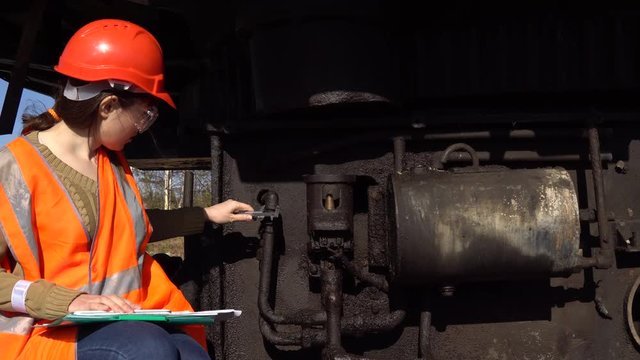 A young woman engineer in an orange waistcoat and a helmet checks the condition of the hydraulic system of a career excavator.