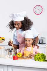 african american mother and daughter in chef hats cooking together on kitchen