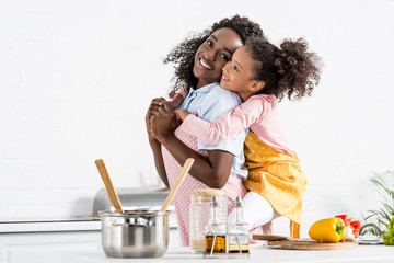 african american mother piggybacking daughter in apron on kitchen