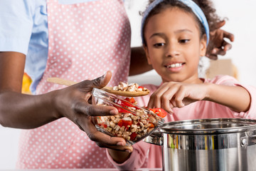 african american mother and daughter putting ingredientes in saucepan on kitchen