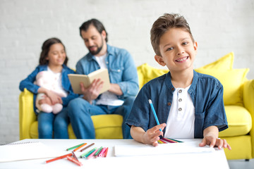 cute little boy drawing with colored pencils and smiling at camera while father and sister sitting on sofa behind