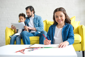 adorable child drawing with colored pencils and smiling at camera while father and brother sitting on sofa behind