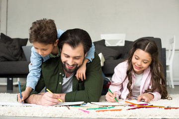 happy father with cute children lying on carpet and drawing with colored pencils