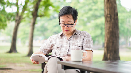 Asian senior woman reading book drinking coffee in sunny park, green background