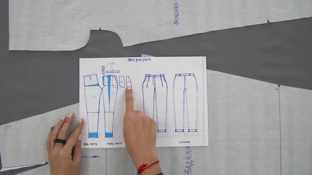 Female hands show something on a pattern for sewing trousers made of gray fabric