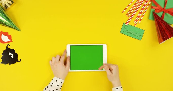 Top view on the white tablet computer on the yellow table with birthday party decorations and writing Surprise while woman's hand scrolling, taping and zooming on the green screen. Horizontally
