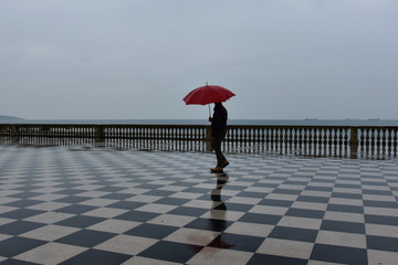 Rainy day in Livorno, Italy. Dark silhouette of a man with red umbrella walking on the wet promenade by the sea.
