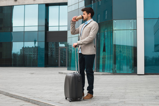 Look well to this day! Full length photo of the handsome delightful entrepreneur drinking delicious coffee and standing with his baggage near the airport, while waiting for the cab.