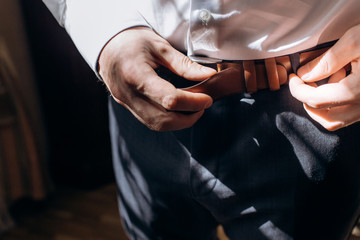 Groom adjusting stylish brown leather belt during morning before wedding, handsome man in elegant suit putting on belt before meeting closeup