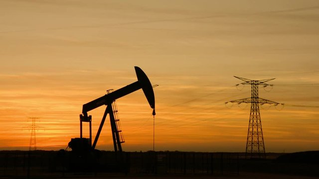 Pumpjack and transmission tower at sunset symbolizing energy transition. Silhouette of a pump jack pumping oil out of a well with an electricity pylon and a power line against a red sky.