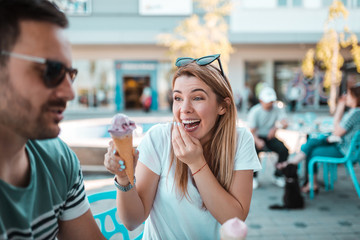 Happy young blonde woman is having ice-cream while she is sitting with her boyfriend outdoors