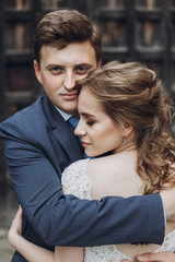Happy couple of newlyweds posing near ancient wooden castle gates, handsome smiling groom embracing beautiful blonde bide in lace wedding dress, family portrait concept