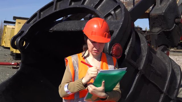 A young woman worker in an orange vest stands near a large ladle of a quarry excavator, looking over project.