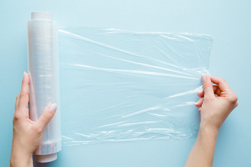 Woman's hand using a roll of transparent polyethylene food film for packing products on the pastel...